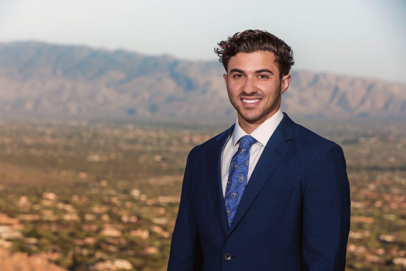 a man wearing a suit and tie standing in front of a mountain